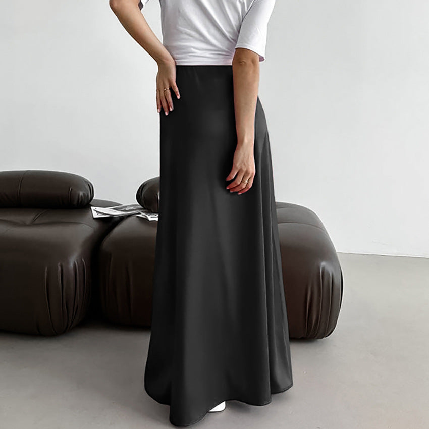 Frau in langem, schwarzem Maxirock und weißem T-Shirt steht in modernem Wohnzimmer mit braunem Sofa. Elegante Mode, minimalistisches Interieur.