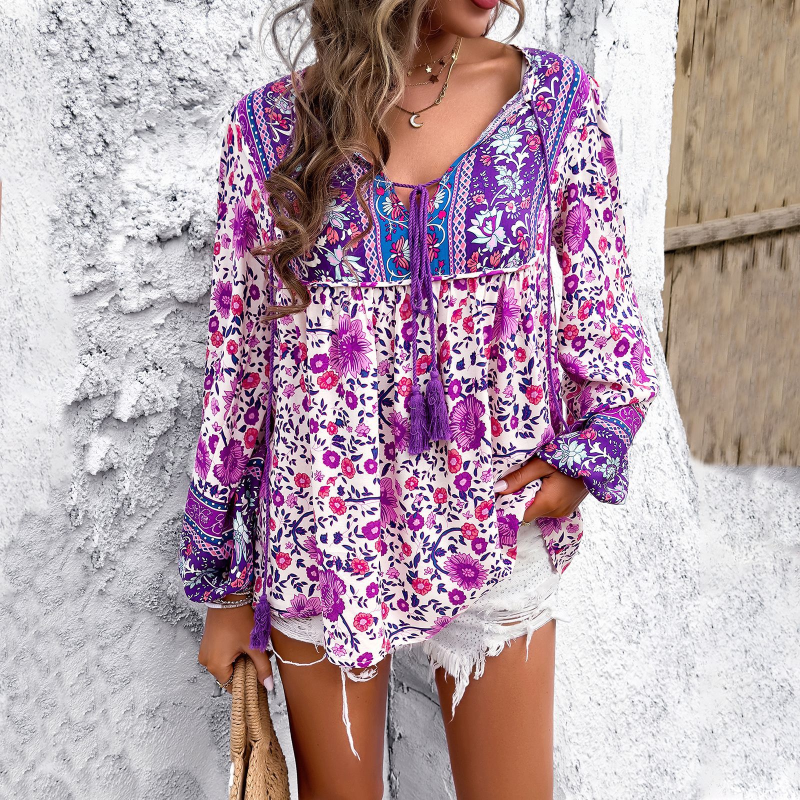 Frau in lila geblümter Boho-Bluse und weißen Shorts vor weißer Wand. Sommermode, Boho-Stil, lässiges Outfit, trendige Damenbekleidung.