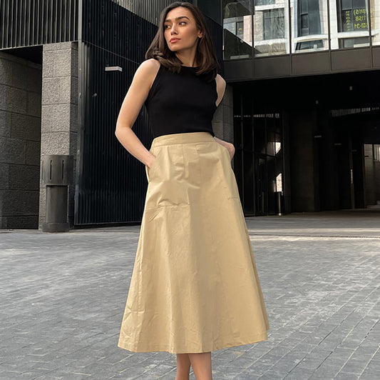 Frau in schwarzem ärmellosen Top und beigem A-Linien-Rock steht vor modernem Gebäude. Elegante Mode, urbaner Stil, Sommeroutfit.