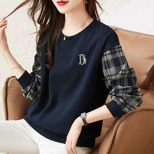 Frau trägt dunkelblauen Pullover mit karierten Ärmeln, sitzt auf einem beigen Sofa. Modische Damenbekleidung, lässiger Stil, Herbstmode.