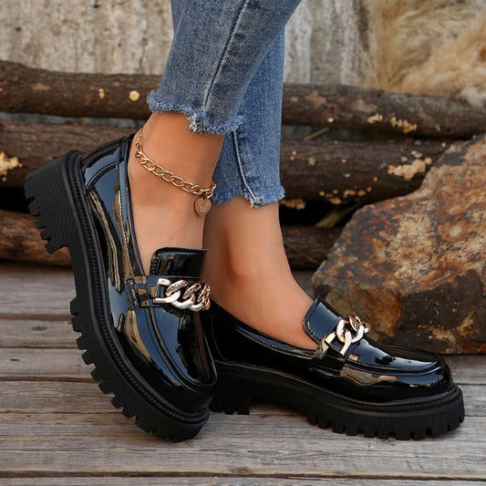 Schwarze Lackleder-Loafer mit goldener Kette, getragen von Person in blauen Jeans. Modische Damen-Schuhe, trendiger Look, Outdoor-Hintergrund.