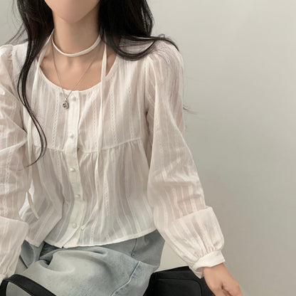 Frau in weißer, langärmeliger Bluse mit Knopfleiste und Halskette, sitzt auf einem Stuhl. Modetrend, elegante Damenbekleidung, lässiger Stil.