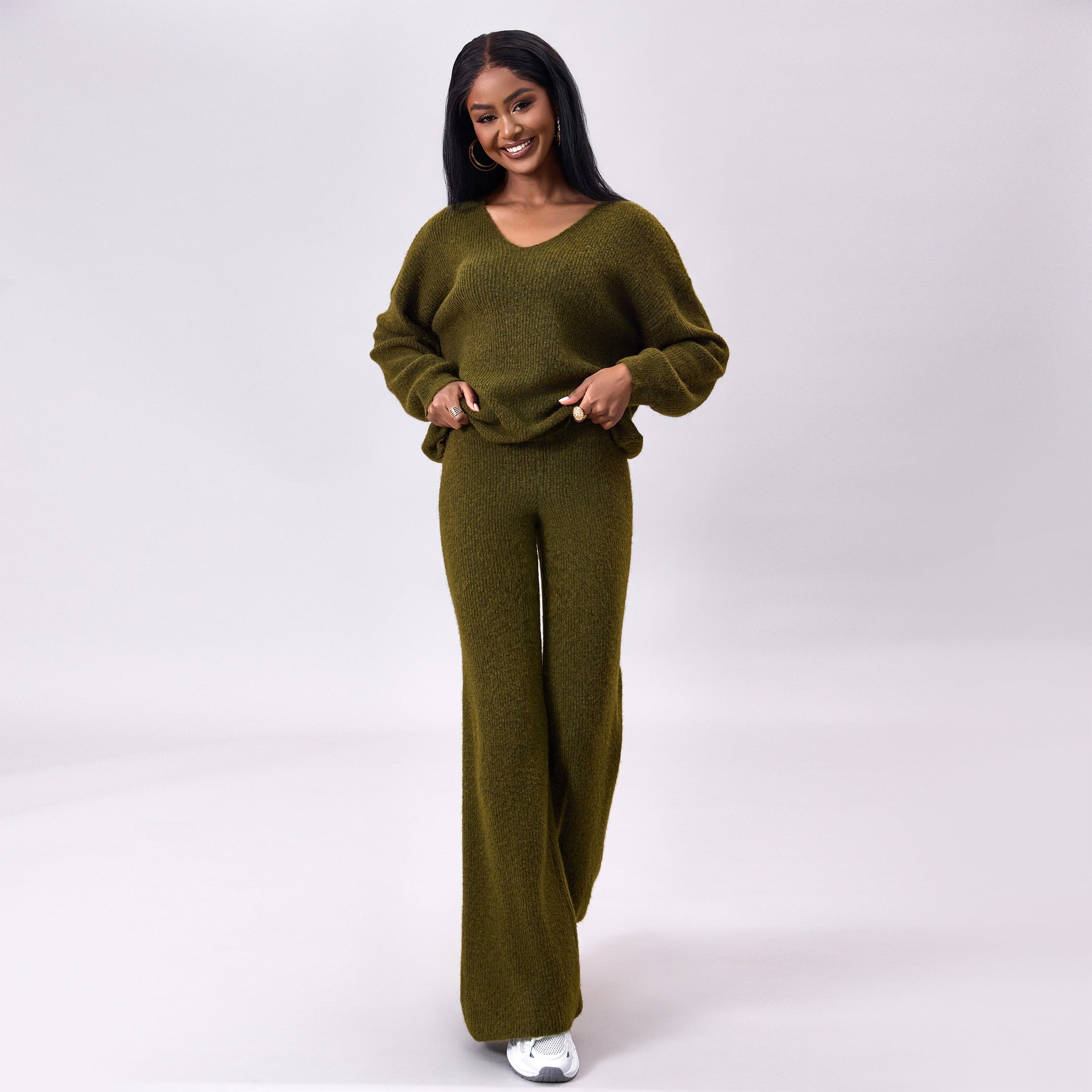 Frau in olivgrünem Strickanzug mit V-Ausschnitt und weiten Hosen, lächelt und steht vor neutralem Hintergrund. Modetrend, Damenbekleidung, Herbstmode.