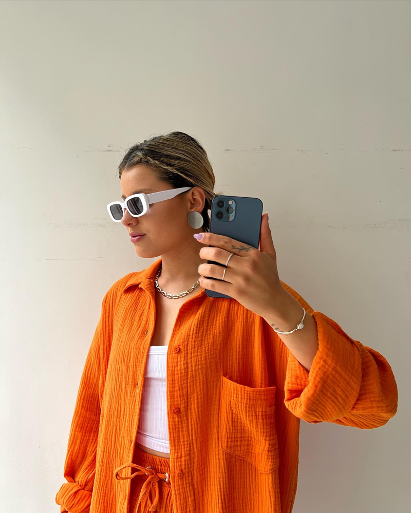 Frau in orangefarbener Bluse mit weißer Sonnenbrille hält ein Smartphone. Trendige Mode, lässiger Stil, moderne Accessoires.