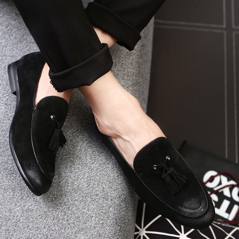 Schwarze Herren-Slipper aus Wildleder mit Quasten, getragen von einer Person auf grauem Sofa. Elegante, modische Schuhe für stilvolle Anlässe.