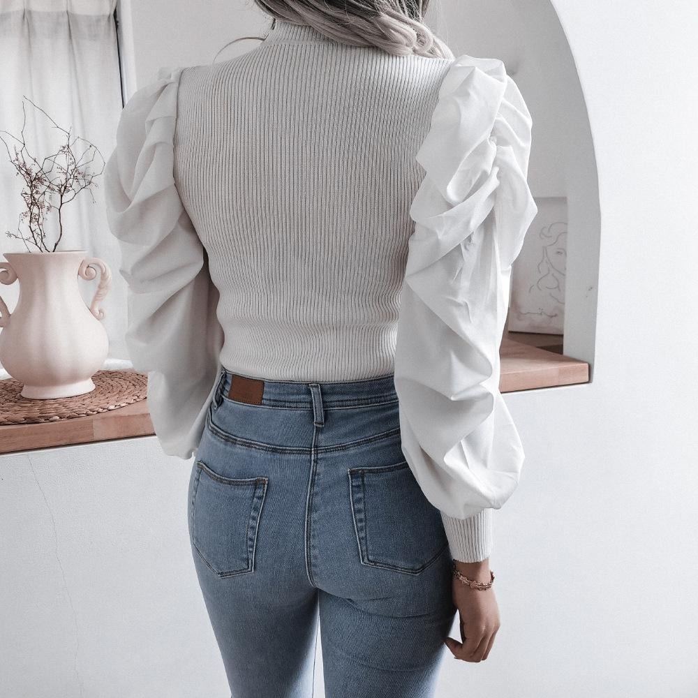 Frau in weißem Pullover mit Puffärmeln und blauer Jeans, steht in modernem Raum. Mode, Damenbekleidung, stilvoll, elegant, Rückansicht.
