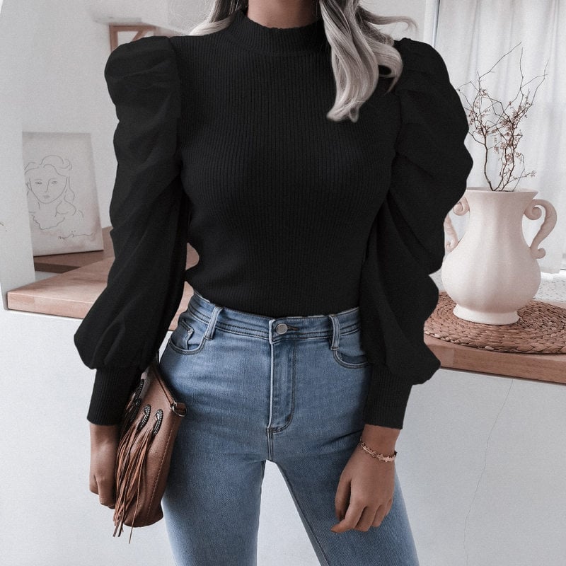 Frau in schwarzem Pullover mit Puffärmeln und blauer Jeans, steht vor einem Tisch mit Vase. Modetrend, eleganter Look, Herbstmode.