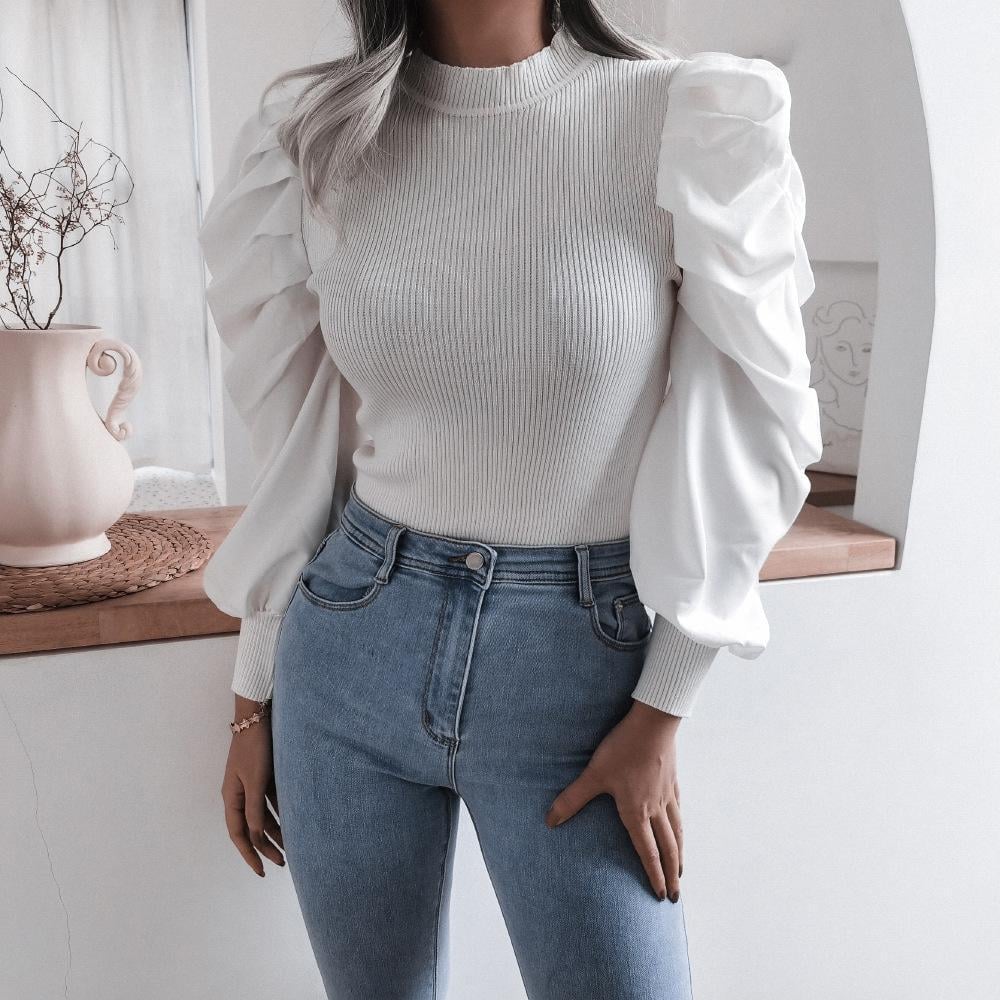 Frau in weißem Pullover mit Puffärmeln und blauer Jeans, steht vor einem Tisch mit Vase. Modetrend, elegante Damenbekleidung, stilvolles Outfit.