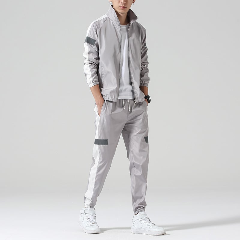 Grauer Herren-Trainingsanzug mit weißen Streifen, bestehend aus Jacke und Jogginghose, kombiniert mit weißen Sneakers. Sportlich, modern, lässig.