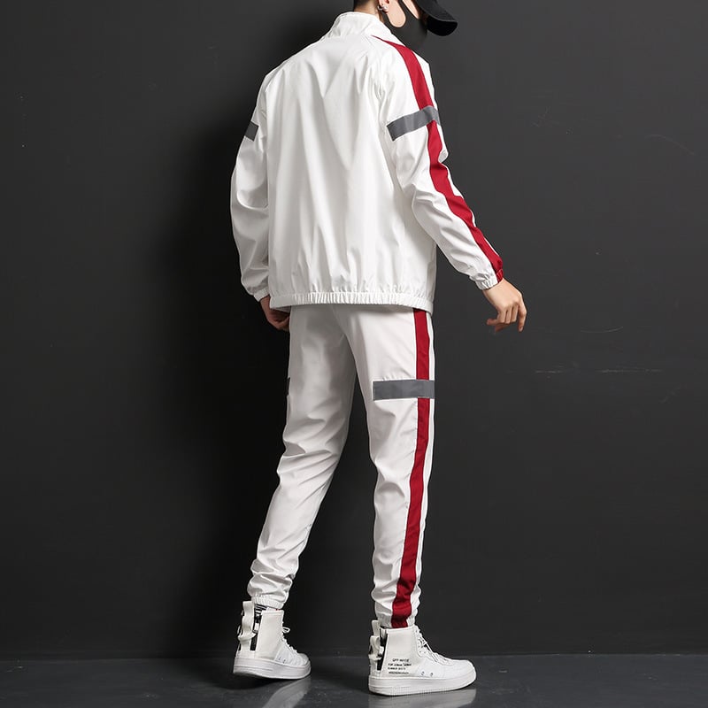 Mann in weißem Trainingsanzug mit roten Streifen, schwarze Mütze, weiße Sneakers, steht vor schwarzem Hintergrund. Sportmode, Streetwear, Herrenbekleidung.