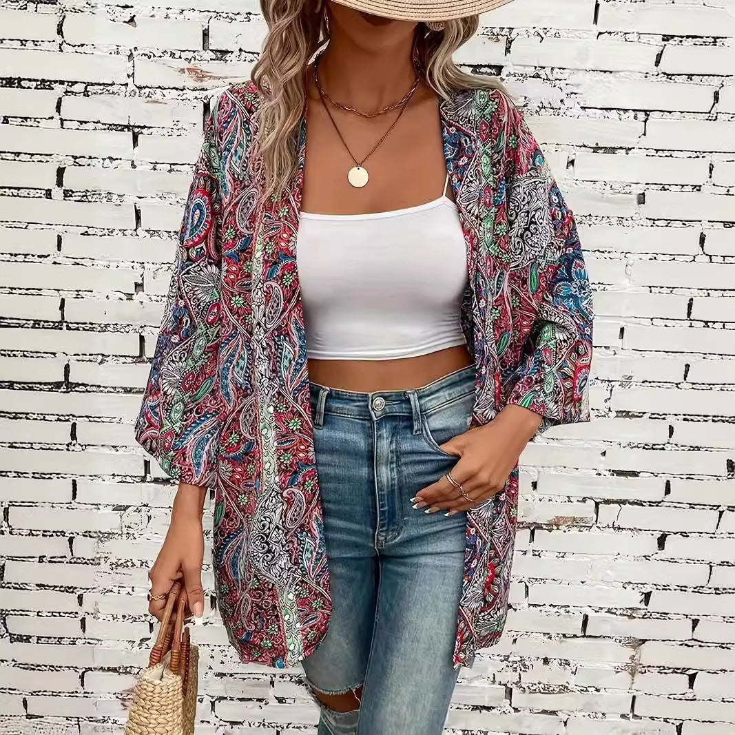 Frau in bunter Paisley-Kimono-Jacke, weißem Crop-Top und blauen Jeans vor weißer Backsteinmauer, trägt Strohhut und geflochtene Tasche.