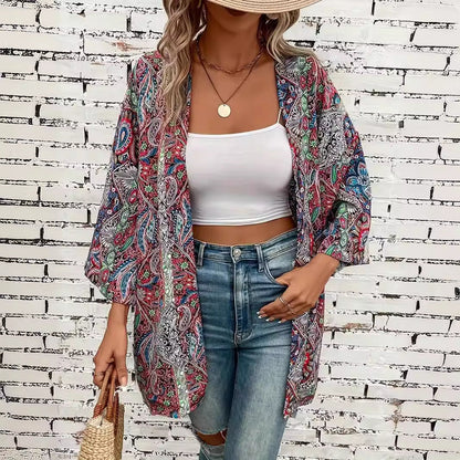 Frau in bunter Paisley-Kimono-Jacke, weißem Crop-Top und blauen Jeans vor weißer Backsteinmauer, trägt Strohhut und geflochtene Tasche.