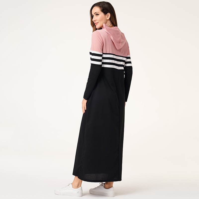 Frau in langem, sportlichem Kapuzenkleid mit rosa und schwarzen Streifen, seitlich stehend, weiße Sneaker, modisch, lässiger Stil.
