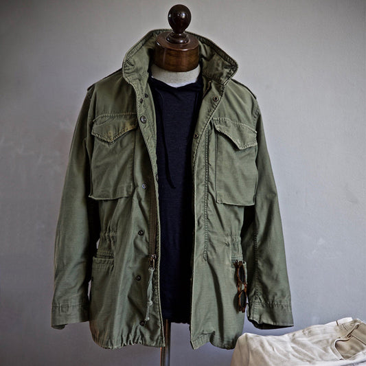 Grüne Militärjacke auf Schneiderpuppe, mit Kapuzenpullover darunter. Outdoor-Mode, Vintage-Stil, robuste Baumwolle, ideal für Herbst und Winter.