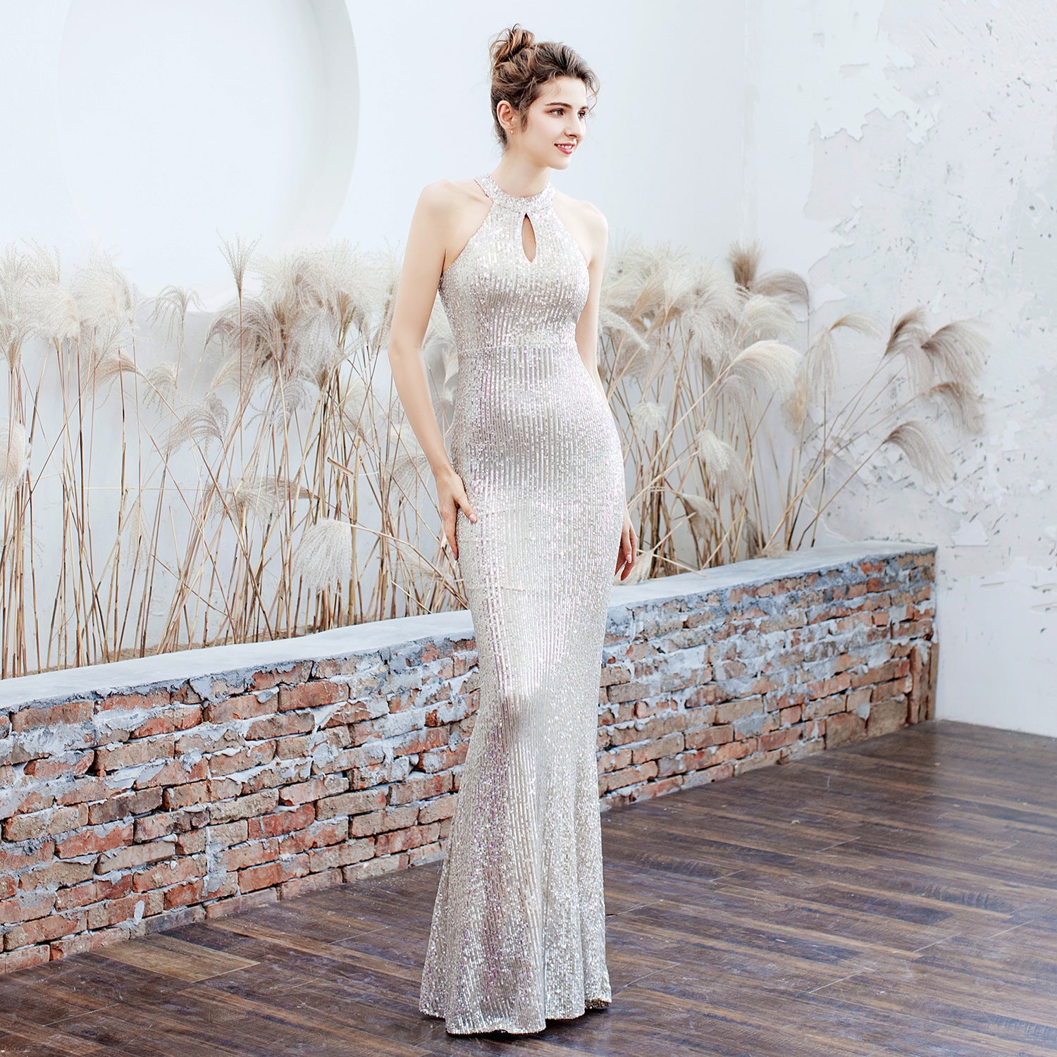 Elegantes Abendkleid in Silber, figurbetont und bodenlang, mit Pailletten und Schlüssellochausschnitt, getragen von einer Frau vor dekorativer Wand.