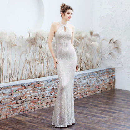 Elegantes Abendkleid in Silber, figurbetont und bodenlang, mit Pailletten und Schlüssellochausschnitt, getragen von einer Frau vor dekorativer Wand.