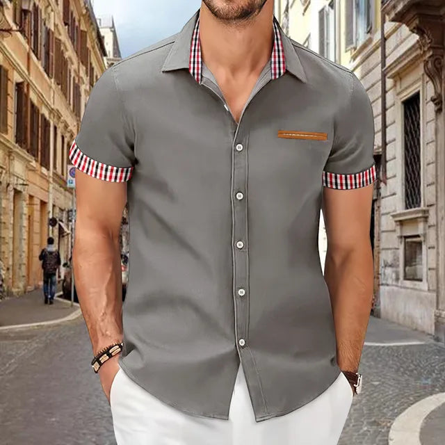 Mann in grauem Kurzarmhemd mit karierten Details, steht auf einer gepflasterten Straße in einer europäischen Stadt. Mode, Herrenbekleidung, urbaner Stil.