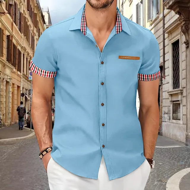 Mann in hellblauem Hemd mit karierten Details, steht auf einer gepflasterten Straße in einer europäischen Stadt. Modetrend, Herrenmode, Sommeroutfit.