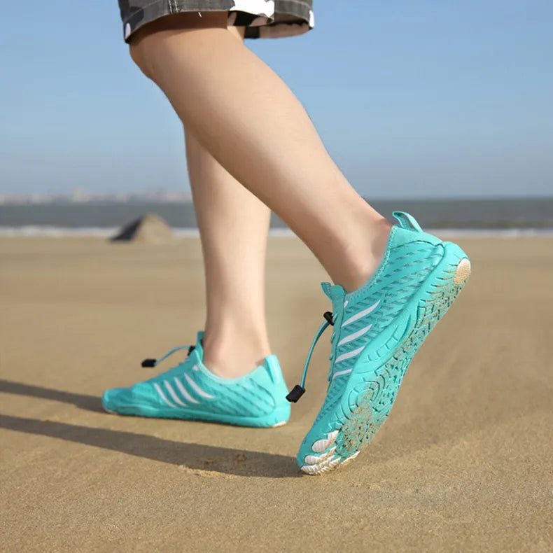 Person in türkisfarbenen Barfußschuhen läuft am Sandstrand entlang. Leichte, flexible Schuhe für Strandspaziergänge und Outdoor-Aktivitäten.