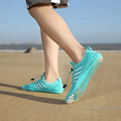 Person in türkisfarbenen Barfußschuhen läuft am Sandstrand entlang. Leichte, flexible Schuhe für Strandspaziergänge und Outdoor-Aktivitäten.