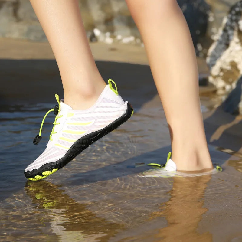 Person trägt weiße Wasserschuhe mit schwarzer Sohle und gelben Akzenten, geht durch flaches Wasser am Strand. Outdoor-Schuhe, Barfußgefühl, Strandwandern.