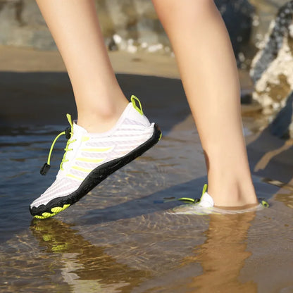 Person trägt weiße Wasserschuhe mit schwarzer Sohle und gelben Akzenten, geht durch flaches Wasser am Strand. Outdoor-Schuhe, Barfußgefühl, Strandwandern.