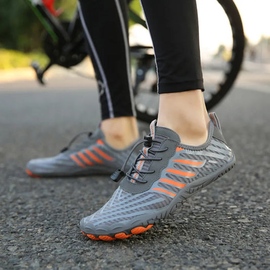 Graue Barfußschuhe mit orangefarbenen Akzenten auf Asphalt, getragen von einer Person in Sportleggings. Ideal für Laufen und Outdoor-Aktivitäten.