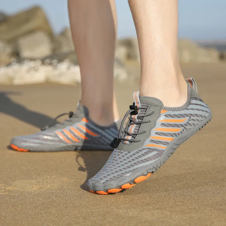 Graue Barfußschuhe mit orangefarbenen Akzenten auf Sandstrand, ideal für Outdoor-Aktivitäten. Leichte, atmungsaktive Schuhe für Sommer und Sport.