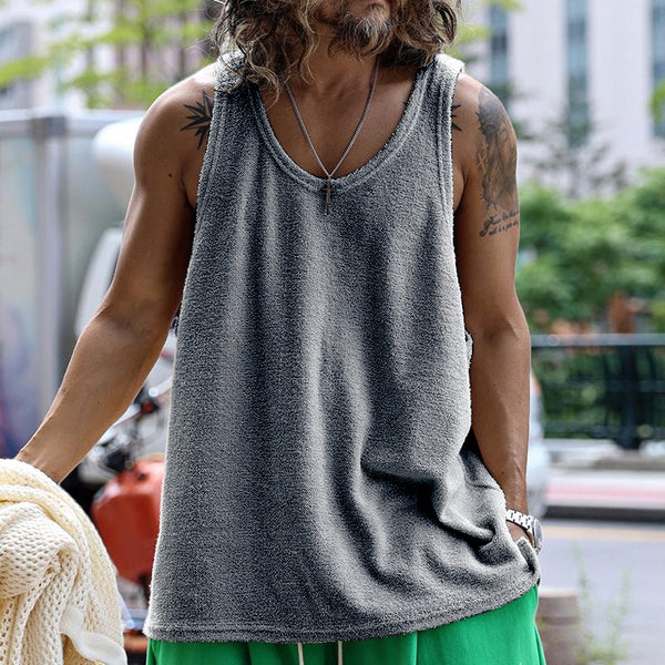 Mann in grauem Tanktop und grüner Hose, trägt Halskette, steht draußen. Sommermode, lässiger Stil, urbaner Hintergrund.