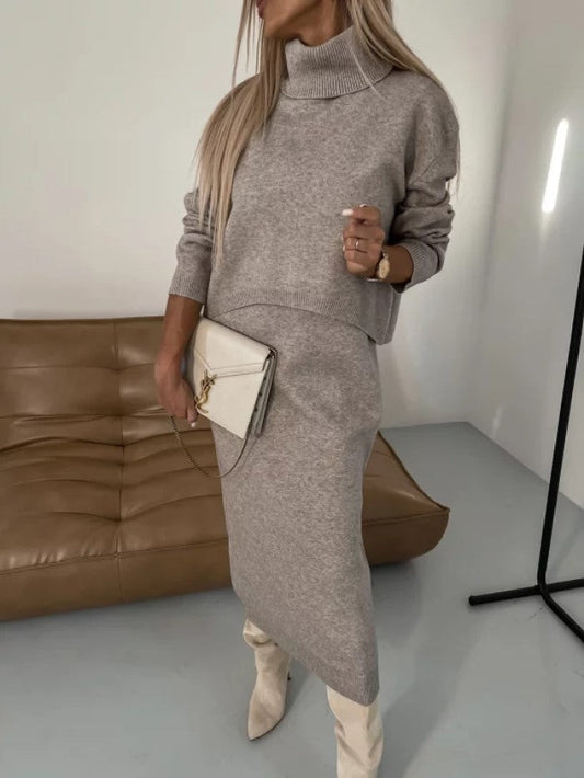 Frau in grauem Strickkleid mit Rollkragen, hält weiße Clutch, steht vor braunem Ledersofa. Modetrend, eleganter Herbstlook, Damenmode.