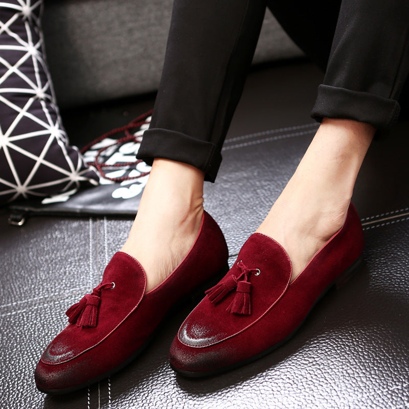 Rote Wildleder-Slipper mit Quasten, getragen von einer Person auf schwarzem Ledersofa. Elegante Herrenschuhe, modisch und bequem.