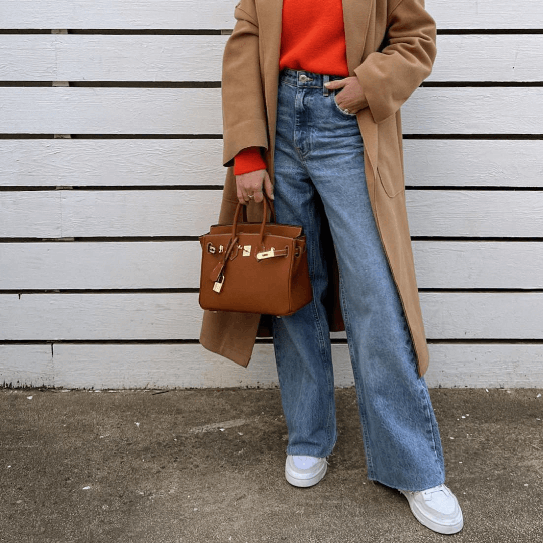 Frau in beigem Mantel und rotem Pullover, trägt blaue Jeans und weiße Sneaker, hält eine braune Designer-Handtasche vor weißem Holzzaun.