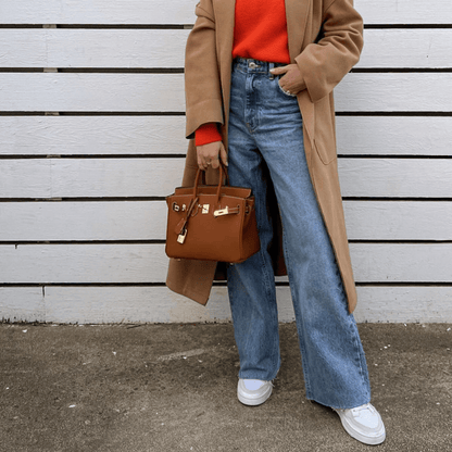 Frau in beigem Mantel und rotem Pullover, trägt blaue Jeans und weiße Sneaker, hält eine braune Designer-Handtasche vor weißem Holzzaun.