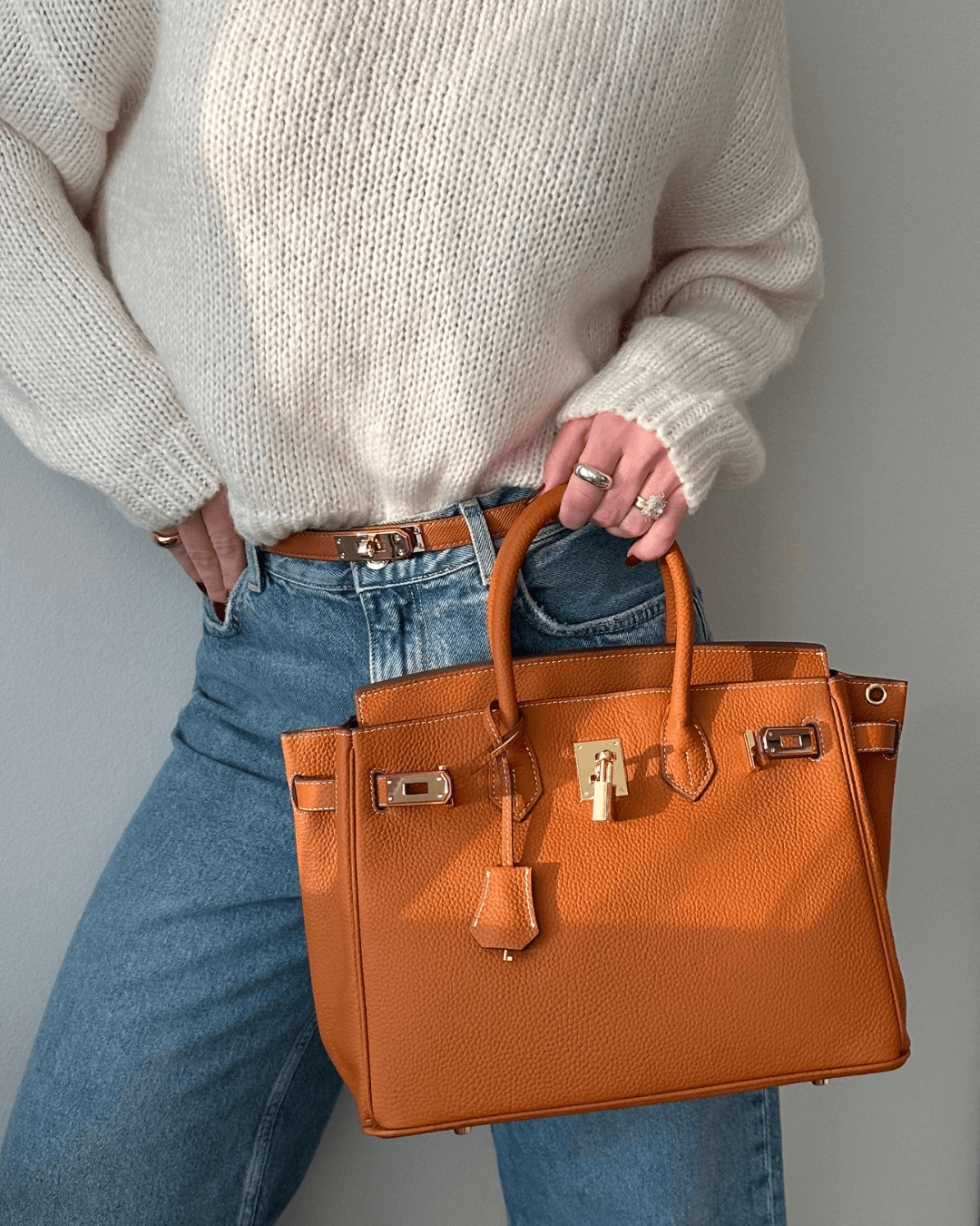 Frau mit weißem Pullover und blauer Jeans hält eine luxuriöse, orangefarbene Lederhandtasche mit goldenen Details. Fashion, Damenmode, Accessoires.