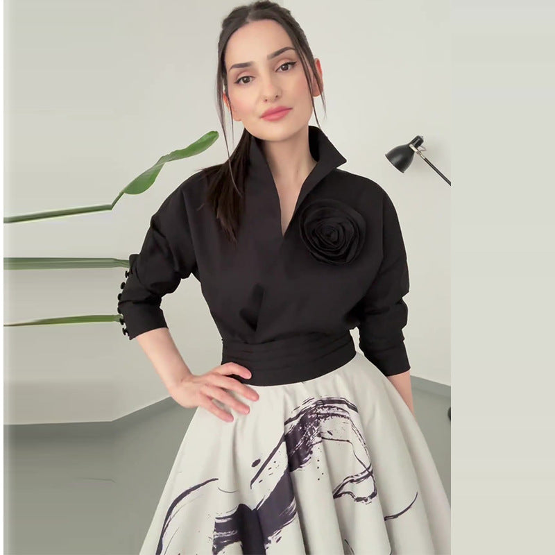Eine Frau in schwarzer Bluse mit Rosenapplikation und weißem Rock mit abstraktem Muster, steht in modernem Raum mit Pflanze und Stehlampe.
