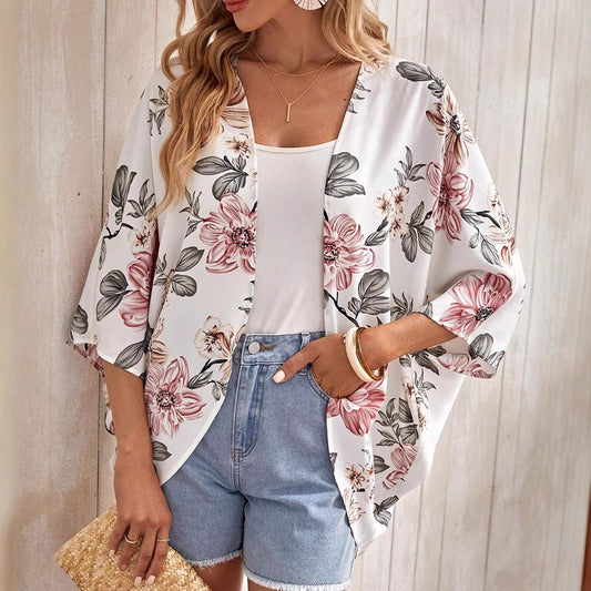 Frau in floralem Kimono, weißem Top und Jeansshorts, hält eine Strohtasche. Sommermode, lässiger Stil, Blumenmuster, Damenbekleidung.