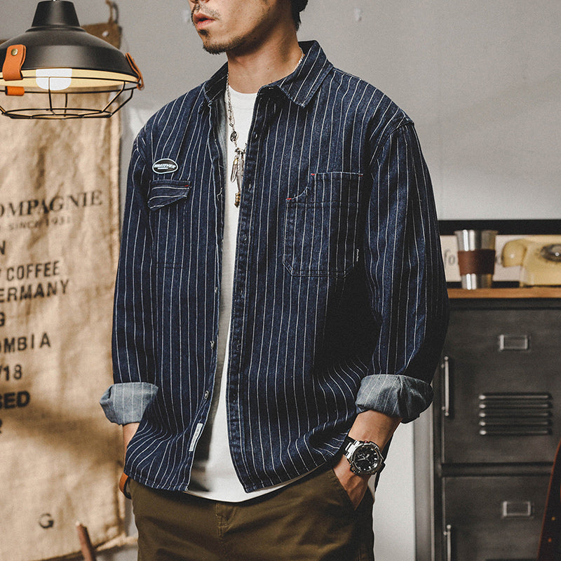Mann trägt gestreiftes Jeanshemd, lässiger Stil, aufgerollte Ärmel, moderne Herrenmode, Vintage-Dekor im Hintergrund, stilvolles Accessoire.