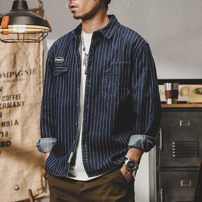 Mann trägt gestreiftes Jeanshemd, lässiger Stil, aufgerollte Ärmel, moderne Herrenmode, Vintage-Dekor im Hintergrund, stilvolles Accessoire.