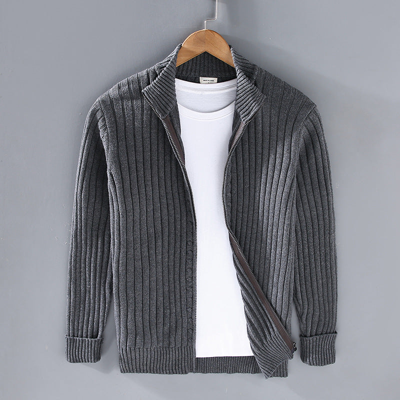 Grauer Strick-Cardigan mit Reißverschluss, über weißem T-Shirt, auf Holzkleiderbügel vor grauer Wand. Herrenmode, stilvoll, lässig.