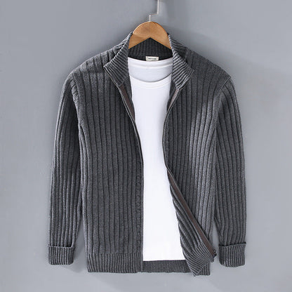 Grauer Strick-Cardigan mit Reißverschluss, über weißem T-Shirt, auf Holzkleiderbügel vor grauer Wand. Herrenmode, stilvoll, lässig.