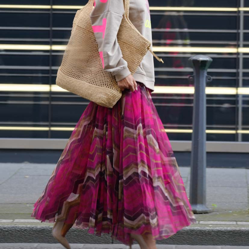 Frau mit beigem Stricktasche und pinkem, gemustertem Maxirock auf Gehweg. Modetrend, Streetstyle, urbaner Look, Herbstmode.
