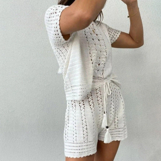 Frau in weißem, gehäkeltem Sommeroutfit mit kurzen Ärmeln und Shorts, posiert vor neutralem Hintergrund. Modetrend, Häkelmode, Sommerkleidung.