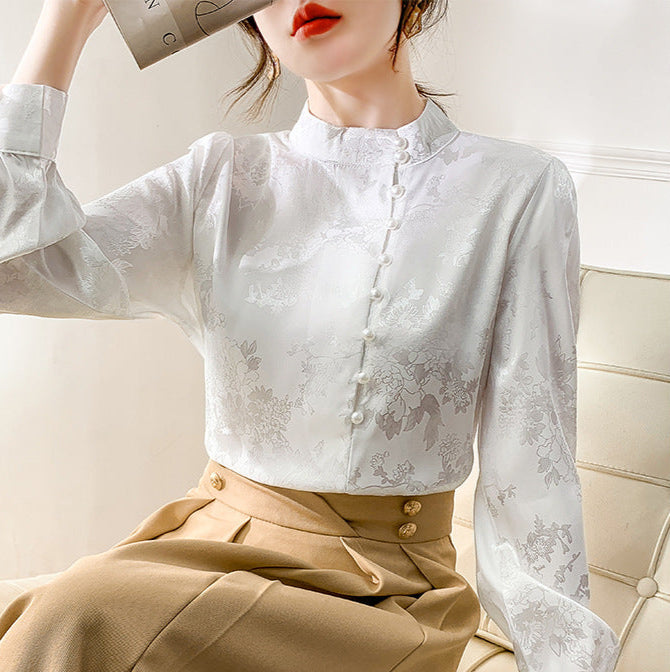 Frau in eleganter weißer Bluse mit floralem Muster, sitzt auf cremefarbenem Sofa, hält Zeitschrift. Modetrend, Damenmode, stilvolle Kleidung.