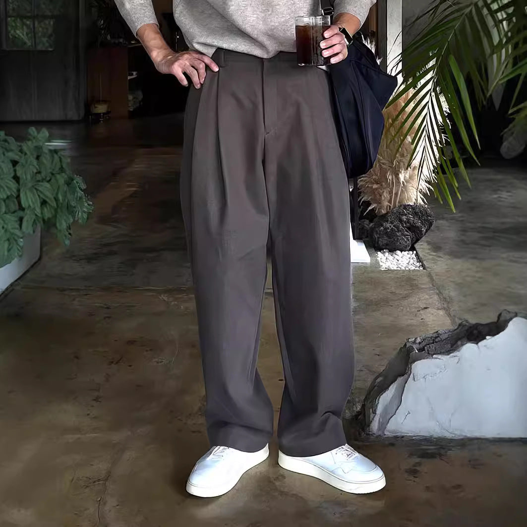 Graue weite Hose, weißes T-Shirt, weiße Sneaker, Person hält Getränk in der Hand, urbaner Hintergrund mit Pflanzen, lässiger Stil.