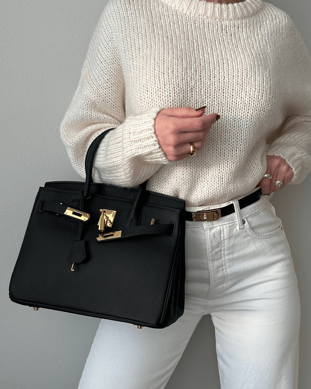 Frau in weißem Strickpullover und Jeans hält eine schwarze Designer-Handtasche mit goldenen Details. Elegante Mode, Luxusaccessoire, stilvoll.