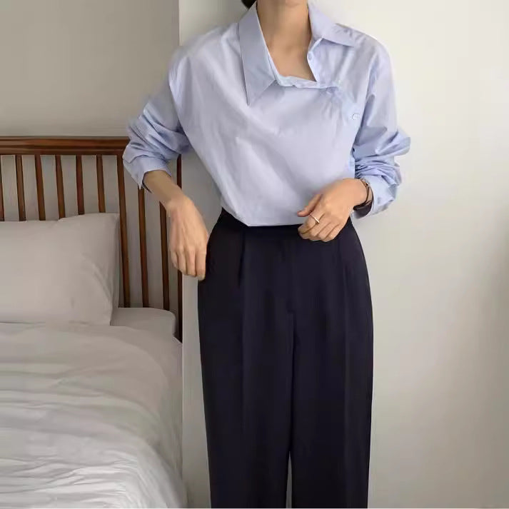 Frau in hellblauer Bluse und schwarzer Hose steht neben einem Bett. Elegante, minimalistische Mode, Schlafzimmer, moderne Kleidung.