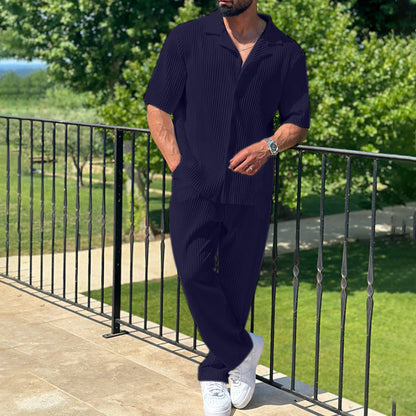 Mann in dunkelblauem Sommeranzug und weißen Sneakers steht auf Balkon mit grünem Garten im Hintergrund. Elegante Herrenmode, Freizeitlook.