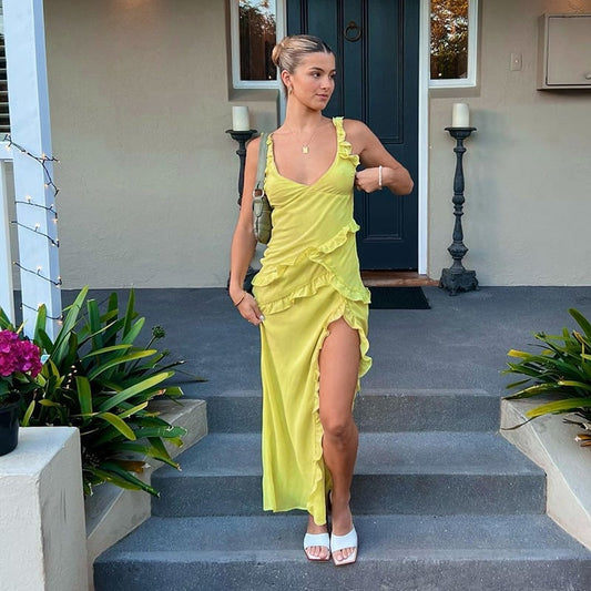 Frau in gelbem Kleid mit Rüschen und hohem Schlitz steht auf Treppe vor Haustür, umgeben von Pflanzen. Elegante Abendmode, Sommerlook.