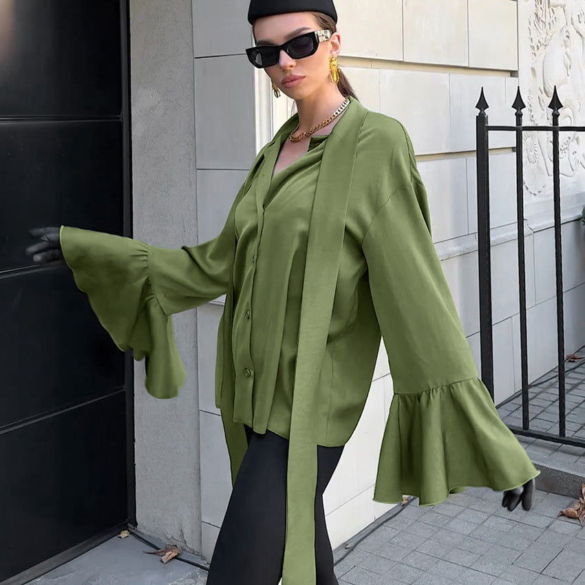 Frau in grüner Bluse mit Glockenärmeln, schwarzer Hose und Sonnenbrille posiert stilvoll vor einem modernen Gebäude. Elegante Mode, urbaner Stil.