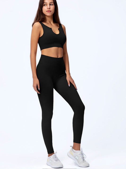 Frau in schwarzem Sport-BH und Leggings, stehend vor weißem Hintergrund, trägt graue Turnschuhe. Fitnessmode, Sportbekleidung, Athleisure.